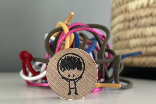 poppetje getekend op houten ronde schijf met daarachter gekleurde veters die in de knoop zitten 